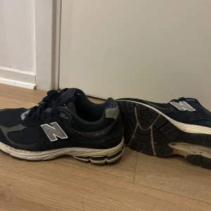 New Balance 2002R mörkblå sneakers - Säljer ett par New Balance 2002R sneakers i mörkblått med grå och vita detaljer. Skorna har mesh och mocka i ovandelen, klassisk N-logga på sidan och snörning framtill. Sulan är vit med svarta och grå inslag, perfekt för dig som gillar retrostil och komfort.
