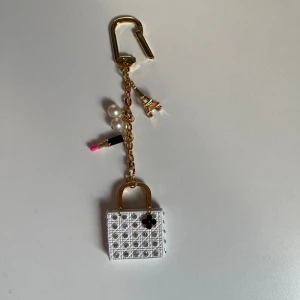 Väska-charm med pärlor och Eiffeltorn - Superfin charm till väska eller nyckelring med detaljer som vit väska med glittriga stenar, guldfärgade handtag, svart blomma, två pärlor, Eiffeltorn och ett läppstift med rosa topp. Kedjan är guldfärgad och ger en lyxig vibe. Perfekt accessoar för att pimpa din väska.