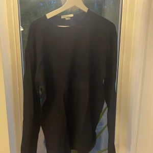 Svart stickad tröja John Smedley L - Snygg svart stickad tröja från John Smedley i storlek L. Klassisk rund halsringning och långärmad modell, perfekt för lager-på-lager. Tillverkad i mjukt material som känns skönt mot huden.