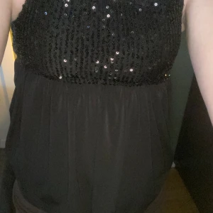 Svart paljettlinne från Gina Tricot - Snygg svart linne från Gina Tricot med tunna axelband och glittriga paljetter upptill. Toppen har en loose passform och är tillverkad i polyester. Perfekt för dig som vill sticka ut med lite extra bling och glam.