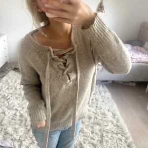 Beige stickad tröja med snörning - Mysig beige stickad tröja med grov snörning framtill och bred ribbad mudd vid ärmslut och nederkant. 