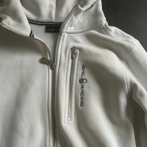 Vit hoodie från Sail Racing xs/164 - Vit hoodie från Sail Racing med dragkedja framtill och på bröstfickan. Tröjan har broderad logga på bröstet och tryck på huvan. Mjuk bomullskänsla, långärmad och med huva för extra chill vibe.