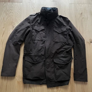 Herno fieldjacka - Säljer en mörkbrun fieldjacka från Herno med fyra stora fickor framtill och hög krage. Jackan är tillverkad i polyester och polyamid, med foder i polyester och bomull. Perfekt för dig som gillar stilren och funktionell design. Är i jättefint skick. Skriv gärna om du undrar något.