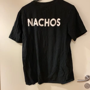 Svart t-shirt med NACHOS-tryck från Saligia - Svart t-shirt från Saligia med klassisk passform och rund hals. På ryggen står det 'NACHOS' i stora vita bokstäver. Tillverkad i mjuk bomull och har korta ärmar. Perfekt för dig som gillar enkla statement-plagg.
