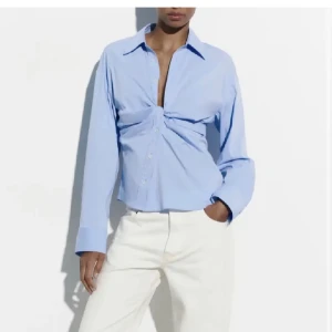 Populär zara skjorta - Snygg ljusblå skjorta med twistad detalj framtill och klassisk krage. Skjortan har långa ärmar, knappar hela vägen och en croppad passform som ger en modern vibe. Perfekt för dig som vill sticka ut med en stilren men unik look.
