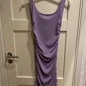 Lila ribbad långklänning från SHEIN - Supersnygg lila långklänning från SHEIN i ribbad stretchig polyester. Klänningen är ärmlös med rundad hals och har snygga rynkade detaljer längs sidan samt knyt längst ner för extra cool look. Perfekt för dig som gillar trendiga och bekväma plagg.