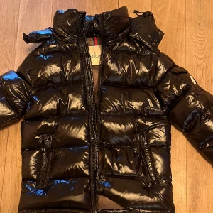 Svart Moncler pufferjacka med huva - Svart glansig pufferjacka från Moncler med huva och dragkedja framtill. Jackan har två fickor med dragkedja och Moncler-logga på ärmen. Den är quiltad och har en riktigt fet streetstil som passar perfekt när det är kallt ute. Använd i 2 dgr,