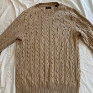 Beige kabelstickad tröja från Gant - Kabelstickad beige tröja från Gant med rund halsringning och diskret logga på bröstet. Tröjan har långa ärmar och ribbade muddar vid ärmslut och nederkant. Perfekt för lager-på-lager och snygg till jeans eller chinos.