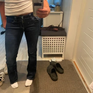 Mörkblå bootcut jeans med kontrastsömmar - Säljer ett par mörkblå lowwaist jeans med bootcut passform och vita kontrastsömmar. Jeansen har klassiska fickor fram och bak, bakfickorna har lock och knapp. 