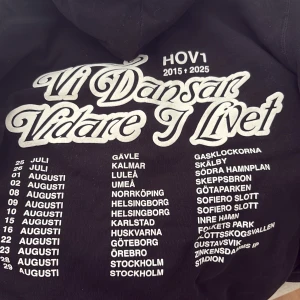 Svart Hov1 hoodie merch 2025 - Svart hoodie från Hov1 med vit kronlogga på bröstet och stort turnétryck på ryggen med texten 'Vi Dansar Vidare I Livet' och turnédatum.  Jag har M och den sitter bra. Pris kan diskuteras 