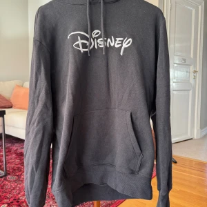 Svart Disney hoodie med tryck - Svart hoodie från Disney med vit logga framtill. Klassisk känguruficka, huva med snörning och långa ärmar. Perfekt för dig som vill ha en chill och snygg streetstil.