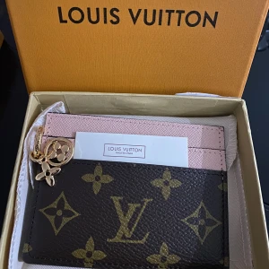 Louis Vuitton plånbok  - Supersnygg plånbok från Louis Vuitton med klassiskt monogrammönster i brunt och guld. Framsidan har LV-logga och blommor, baksidan är ljusrosa. Plånboken har guldfärgade detaljer och charm, perfekt för kort och cash❤️