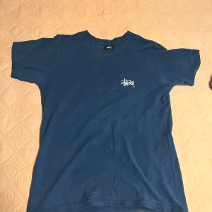 Blå Stüssy t-shirt med stor logga - Säljer en blå t-shirt från Stüssy i 100% bomull. T-shirten har korta ärmar, rund hals och en liten Stüssy-logga på bröstet samt en stor vit Stüssy-logga på ryggen. Perfekt för dig som gillar streetwear och vill sticka ut med en klassisk look.