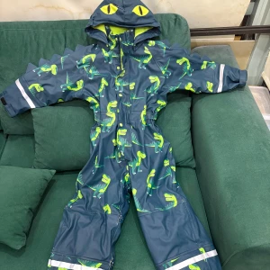 Blå regnoverall med dinosaurier från Ellos - Säljer en blå regnoverall från Ellos med grönt fleecefoder och coolt dinosauriemönster. Overallen har huva med ögon, taggar längs ryggen och reflexdetaljer på ärmar och ben. Perfekt för dig som gillar att sticka ut i regnet!