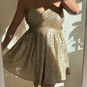 Guldig paljettklänning axelbandslös - Superfin axelbandslös kort klänning i guld med massor av paljetter. Klänningen har en hjärtformad byst, markerad midja och vippig kjol med tyll under. Perfekt för dig som vill sticka ut och glänsa. Dragkedja i ryggen för enkel påklädning. - Använd en gång. 