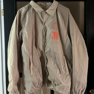 Beige vindjacka Urban Outfitters L - Snygg beige vindjacka från Urban Outfitters med orange grafiskt tryck på bröstet och ryggen. Jackan har krage, tryckknappar framtill och två fickor. Materialet är lätt och syntetiskt, perfekt för lager-på-lager. Passformen är loose och jackan är långärmad. Är lite mindre i storlek, hade kunnat passa Strl.M också