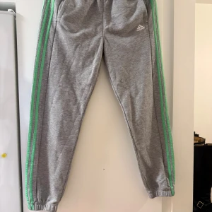 Grå Adidas mjukisbyxor med gröna ränder - Grå mjukisbyxor från Adidas med gröna ränder längs sidorna och klassisk logga på låret. Byxorna har resår i midjan och vid bensluten, samt är tillverkade i ett mjukt och bekvämt material. Perfekta för chill eller träning.
