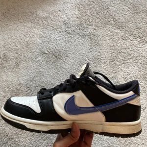 Nike Dunk Low svart/vit/blå - Nike Dunk Low sneakers i svart, vit och blått med klassisk blå Swoosh på sidan. Skorna har snörning, rund tå och platt sula. Ovandelen är i skinn och syntet, med svart meshfoder för extra komfort. Perfekt för dig som gillar streetwear och sportig stil.