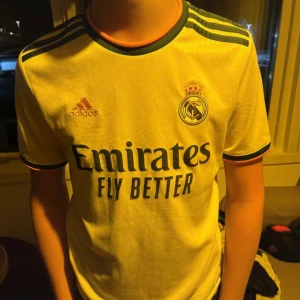 Real Madrid Camavinga 12 Adidas tröja - Vitt Gul Real Madrid fotbollströja från Adidas med Camavinga och nummer 12 på ryggen. Tröjan har svarta detaljer, klubbmärke på bröstet, Adidas-logga och sponsortryck. Klassiska tre ränder på axlarna och rund hals. Tillverkad i lätt funktionsmaterial.