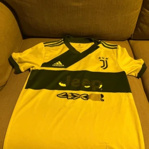 Juventus gul matchtröja Adidas XS - Juventus fotbollströja från Adidas i gul med mörkgrön bred rand och detaljer. Klassisk V-ringning, korta ärmar och klubbmärke på bröstet. Tillverkad i Primegreen-material för en sportig känsla. Snygga kontrastdetaljer på axlar och ärmslut.