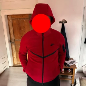 Röd Nike hoodiejacka med dragkedja - Säljer en röd hoodiejacka från Nike med svart logga på bröstet och svarta detaljer längs dragkedjan och på ärmen. Jackan har huva, hel dragkedja och en ficka med dragkedja på vänster ärm. Perfekt för dig som gillar sportig och clean stil. Helt ny