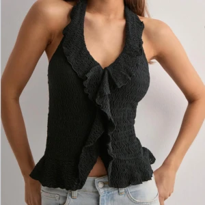 Svart halterneck topp med volang - Supersnygg svart halterneck topp med volangkant och vågig struktur. Toppen knyts i nacken och har en omlottliknande framsida med volangdetaljer längs urringningen. Perfekt för att lyfta vilken outfit som helst.