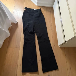 Svarta cut out kostymbyxor NA-KD - Svarta kostymbyxor från NA-KD med trendiga cut out-detaljer i midjan och dragkedja bak. Byxorna har raka ben med lätt utsvängd bootcut och hög midja. Perfekta för dig som vill sticka ut med en modern twist på klassiska byxor.