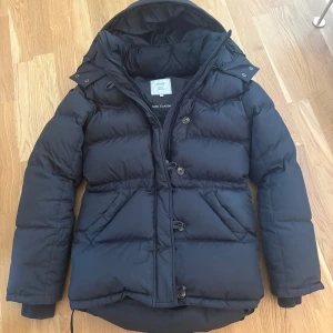 Svart dunjacka från Boomerang XXS - Säljer en svart dunjacka från Boomerang i storlek XXS. Jackan har huva, dragkedja och knappar framtill samt två stora fickor. Klassisk pufferstil med ribbade muddar och justerbar dragsko nertill. Perfekt för kalla dagar.