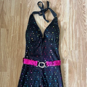 Svart glittrig 70s discodräkt jumpsuit - Svart jumpsuit med halterneck och färgglada glittriga prickar över hela tyget. Klänningen har en djup v-ringning och ett brett, rosa paljettbälte i midjan. Utsvängda ben ger en cool retrovibe. Perfekt för dig som vill klä ut dig till 70 tal. Helt ny och oanvänd, storlek xxs men den är stretchig och passar även en xs/s. Nypris inklusive frakt ca 900 kr för mig.