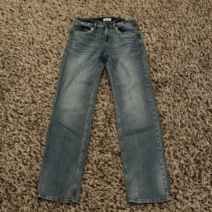 Ljusblå jeans från Lindex, strl 170 - Säljer ett par ljusblå jeans från Lindex i storlek 170. De har klassisk femficksdesign, rak passform och normal midja. Jeansen är tillverkade i bomull och har en snygg tvättad look med subtila slitningar. Perfekt för dig som gillar enkel och stilren denim.