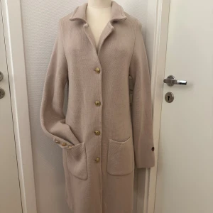 Puderrosa kappa från Busnel - Stilren puderrosa kappa  från Busnel med guldiga knappar med ankardetalj, två stora fickor framtill och klassisk krage. Jackan är stickad i ull och har lång ärm med Busnels ikoniska logga på ärmslutet. Perfekt skick. Storleken är som 38/40 med långa ärmar. 