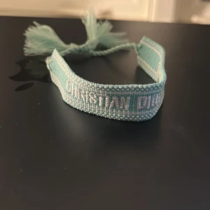 Turkost Christian Dior J'Adior armband - Snyggt turkost armband från Christian Dior med texten 'J'Adior' broderad i vitt. Armbandet är gjort i ett mjukt tygmaterial och har flätade snören med tofsar i ändarna för att knyta runt handleden. Perfekt accessoar för att lyfta din stil.