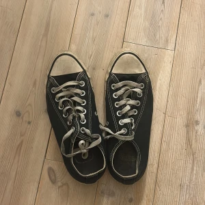 Svarta Converse All Star sneakers - Klassiska svarta Converse All Star sneakers med vit sula och vita snören. Skorna har rund tå, platt sula och tydliga kontrastsömmar. Tillverkade i canvas med gummisula och ikonisk logga på plösen. Perfekta för en avslappnad och cool stil.