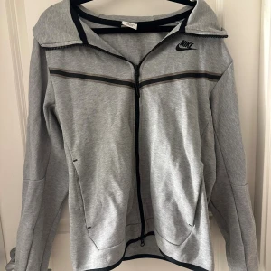 Grå hoodie från Nike med dragkedja - Snygg grå hoodie från Nike med svart logga på bröstet och hel dragkedja framtill. Tröjan har huva, långa ärmar och svarta detaljer längs dragkedjan och muddar. Perfekt för chill eller träning, med två fickor framtill och stilren design.