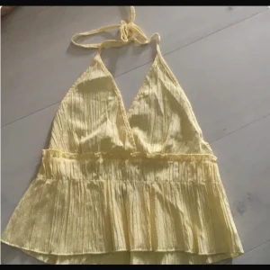 Gul omlott linnetopp med knyt - Säljer en ljusgul omlott linnetopp med knyt i nacken och v-ringad hals. Toppen har volang nedtill och är tillverkad i ett lätt, ribbat material som ger en skön sommarkänsla. Perfekt för dig som gillar en feminin och luftig stil.