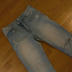 Ljusblå raka jeans med hål på knät - Säljer ett par ljusblå jeans med raka ben och slitningar, bland annat ett stort hål på ena knät. Jeansen har klassisk femficksmodell och är i mjuk denim. Perfekta för en avslappnad och trendig look.