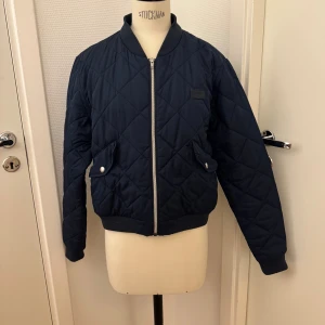 Marinblå bomberjacka från Marqy - Quiltad bomberjacka från Marqy i marinblått med silvrig dragkedja och ribbade muddar. Jackan har två fickor med lock och tryckknappar samt ett diskret märke på bröstet. Tillverkad i 100% polyester. Hämtas i Hägersten om du vill slippa frakt. Var även uppmärksam på när Plick erbjuder fraktfritt ibland på dyrare plagg. Följ mig gärna. Har mycket att lägga ut i olika storlekar! 