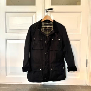 Svart vaxad Fieldjacket Hackett London - Svart Field jacket från Hackett London, modell McGregor Coat. Jackan har fyra stora fickor framtill, knappar och dragkedja samt krage och muddar i manchester. Insidan är fodrad med klassiskt rutigt tyg i grönt, beige och svart.  Självklart helt fläckfri! Perfekt för höst och vinter. Skärp finns.