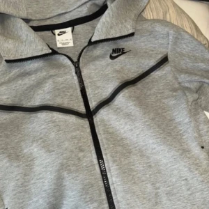 Grå Nike zip-hoodie XS - Snygg grå zip-hoodie från Nike med svarta detaljer och dragkedja hela vägen. Jackan har huva, svarta linjer över bröstet och ärmen samt praktiska fickor. Tillverkad i mjuk bomulls- och polyestermix, perfekt för en sportig och chill stil.