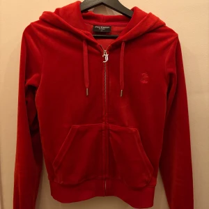 Röd velour hoodie från Juicy Couture - Juicy Couture hoodie i röd velour med dragkedja, huva och snörning. Klassisk JC-logga broderad på bröstet och känguruficka framtill. Perfekt för dig som vill ha en ikonisk och mjuk streetstyle-look.