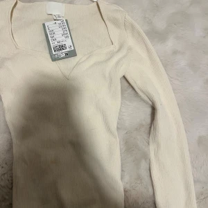 Ribbad vit långärmad topp H&M - Säljer en ribbad vit långärmad topp från H&M med snygg v-ringning och figurnära passform. Toppen har en modern fyrkantig urringning och är perfekt att styla med jeans eller kjol för en trendig look.