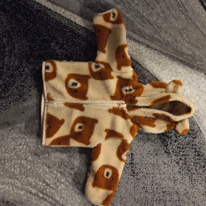 Teddyjacka med björnmönster från Lupilu - Supergosig teddyjacka från Lupilu med huva och dragkedja. Jackan är beige med brunt björnmönster och har fluffig fleeceyta som känns mjuk mot huden. Perfekt för kyliga dagar när du vill vara både söt och varm.