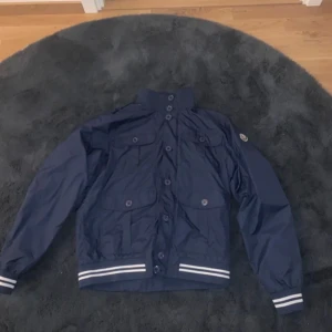 Mörkblå vindjacka från Moncler - Snygg mörkblå vindjacka från Moncler med klassisk logga på ärmen. Jackan har flera fickor med knappar, hög krage och vita ränder på muddar och midja. Tillverkad i lätt polyester, perfekt för dig som vill ha en stilren och sportig look.