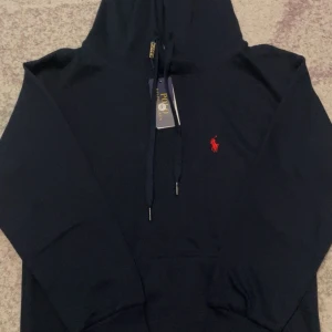 Mörkblå hoodie från Polo Ralph Lauren - Snygg mörkblå hoodie från Polo Ralph Lauren med klassisk röd broderad logga på bröstet. Tröjan har huva med snörning och en stor magficka. Perfekt för dig som gillar stilrena och tidlösa plagg.