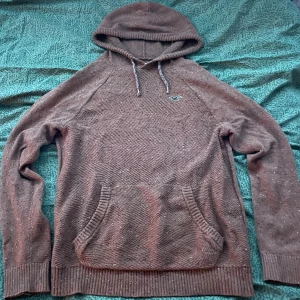 Brun stickad hoodie från Hollister - Mysig brun stickad hoodie från Hollister med huva och dragsnören. Tröjan har ett diskret mönster, magficka och Hollisters klassiska logga broderad på bröstet. Perfekt för kyliga dagar och enkel att matcha med jeans eller joggers.