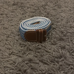 Blått och vitt flätat bälte - Snyggt flätat bälte i blått och vitt med bruna skinn-detaljer och silverfärgat spänne. Bältet har ett coolt mönster och passar perfekt till jeans eller chinos för en avslappnad men stilren look.