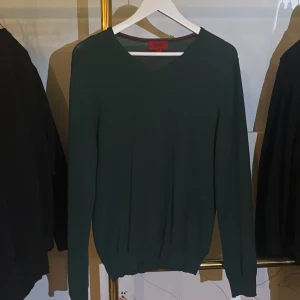 Mörkgrön stickad tröja från Hugo Boss - Snygg mörkgrön stickad tröja från Hugo Boss med v-ringad hals och ribbade muddar. Enkel och stilren design som passar perfekt till jeans eller chinos. Tröjan har lång ärm och är tillverkad i ett mjukt material som känns skönt mot huden.
