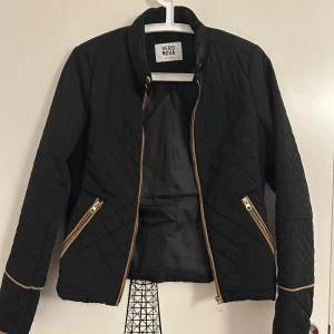 Svart quiltad jacka från Vero Moda - Svart quiltad jacka med guldiga dragkedjor och detaljer i storlek S från Vero Moda. Jackan har lång ärm, ståkrage och två snedställda fickor framtill. Jackan är i bra skick. 