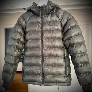 Grå dunjacka Peak Performance - Snygg grå dunjacka från Peak Performance med huva och dragkedja framtill. Jackan har quiltad design, ribbade muddar och två sidofickor med dragkedja. Perfekt för kalla dagar och har en stilren look som funkar till det mesta.