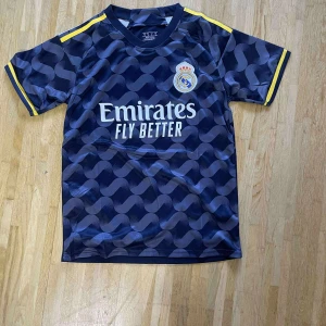 Real Madrid marinblå fotbollströja - Snygg marinblå fotbollströja från Real Madrid med korta ärmar och gul detalj på ärmsluten. Tröjan har ett vågigt mönster på framsidan och klubbens emblem på bröstet. Materialet är lätt och andas, perfekt för träning eller match.